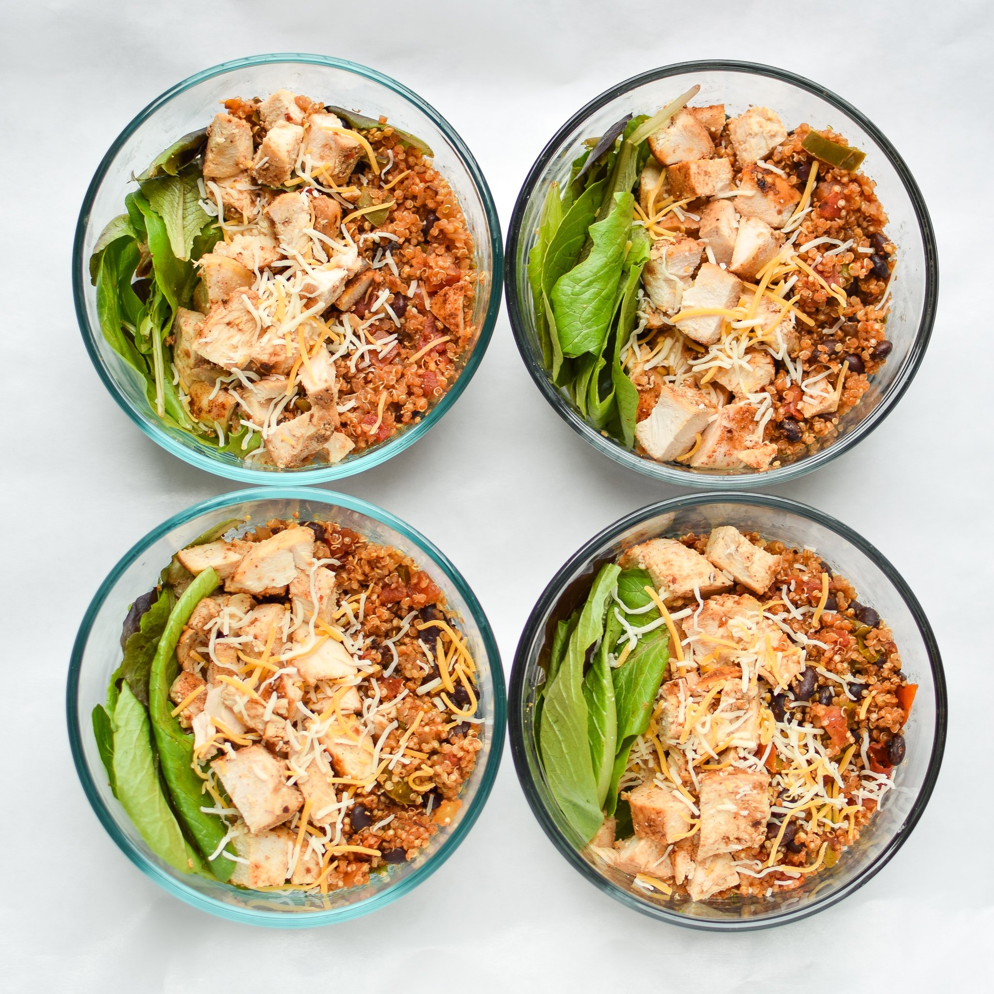 Mexican quinoa chicken salad lunch bowls viewed from above. Chicken breast, mexican quinoa and greens with cheese sprinkled on top in four Pyrex bowls.