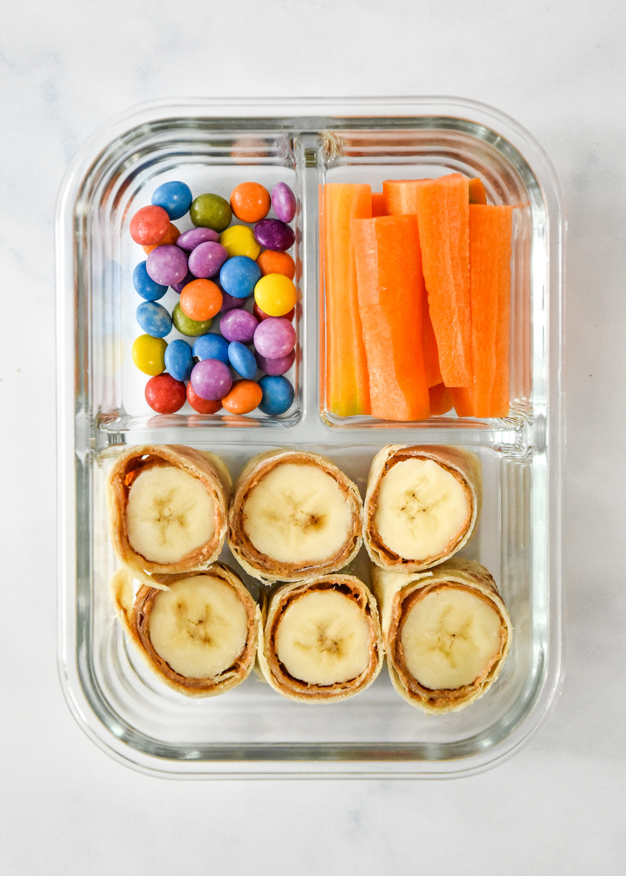 pb banana roll ups with carrot sticks and chocolate candies in a glass meal prep container.
