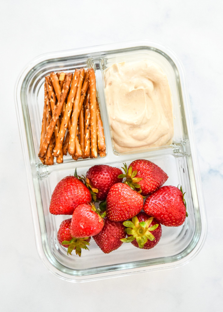 peanut butter greek yogurt fruit dip in a glass meal prep container with strawberries and pretzels.