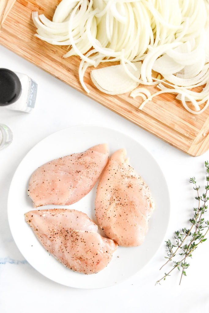 seasoned chicken breasts on a white plate and onions on a cutting board.