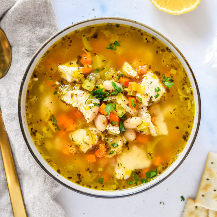 white bean lemon chicken soup in a bowl with veggies and broth, crackers and lemon on the side.