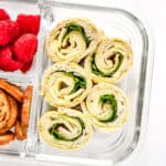 turkey pesto pinwheels in a glass meal prep container with raspberries and bagel chips.
