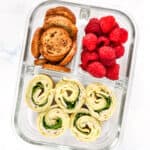 turkey pesto pinwheels in a glass meal prep container with raspberries and bagel chips.